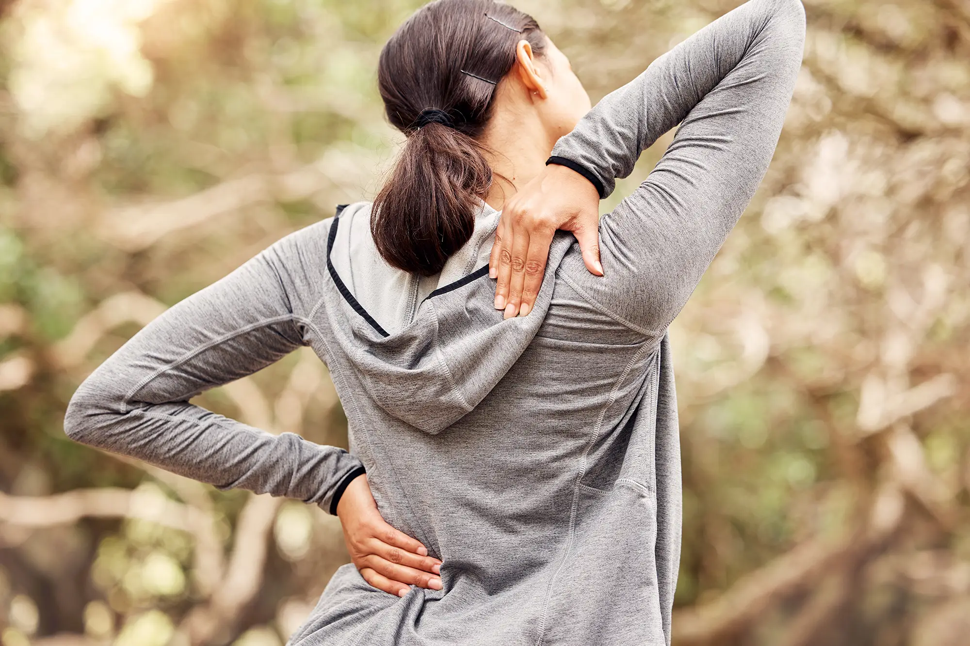 Woman in gray athletic wear experiencing back pain, holding lower back, surrounded by natural setting, illustrating symptoms of back pain and sciatica.