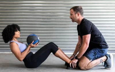 Woman performing sit-up with medicine ball, assisted by personal trainer, in a fitness setting, emphasizing physical therapy and training services.